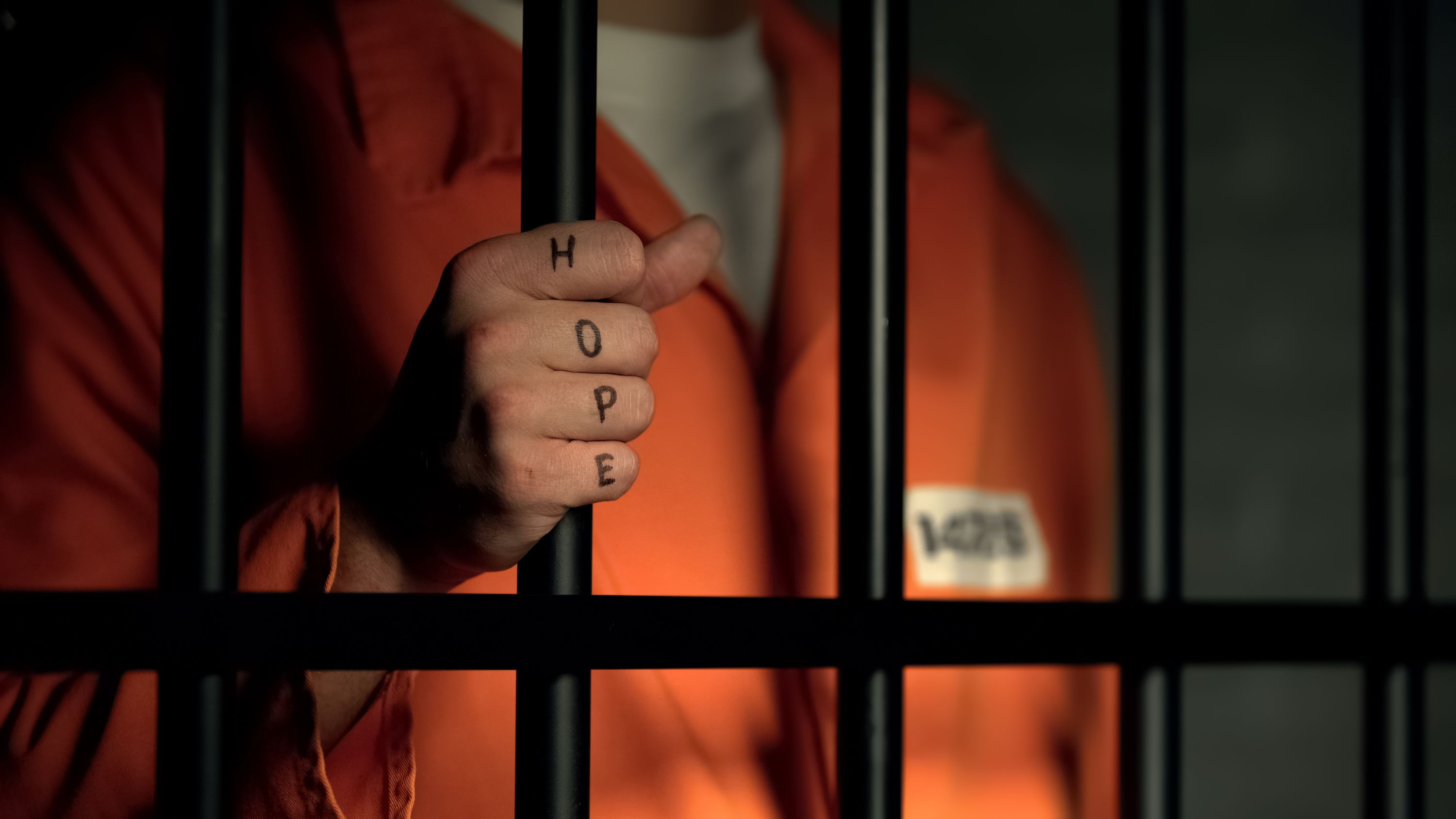 Image: Person in orange with one hand on prison bars, showing letters spelling out H-O-P-E on their knuckles